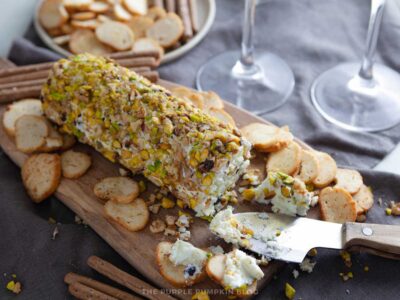 Pistachio and blackcurrant cheese log on a wooden board with crackers, a cheese knife, and wine glasses in the background.