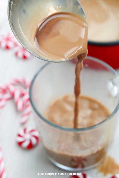 A ladle pours hot chocolate into a glass, with candy canes and peppermint candies blurred in the background on a light wooden surface.