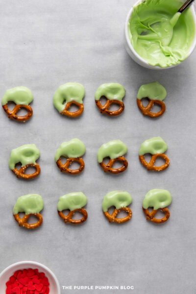 Mini pretzel twists dipped in green candy coating arranged in rows on parchment paper, with a bowl of melted green candy and a bowl of red heart sprinkles nearby.