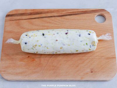 Uncoated pistachio and blackcurrant cheese log wrapped tightly in plastic wrap on a wooden cutting board.