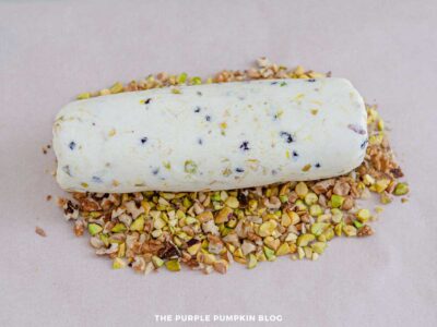 Uncoated cheese log being rolled in a mixture of chopped pistachios and walnuts on parchment paper.