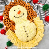 Close-up of a Snowman Cheeseball covered in shredded white cheddar cheese with peppercorn eyes, mouth, and buttons and a baby carrot nose, surrounded by pretzels and crackers on a serving platter.