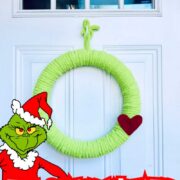 Green yarn Grinch wreath hanging on a white front door with a red felt heart accent, displayed as a holiday decoration. Text overlay says: "The Grinch Wreath Easy Holiday Craft!"
