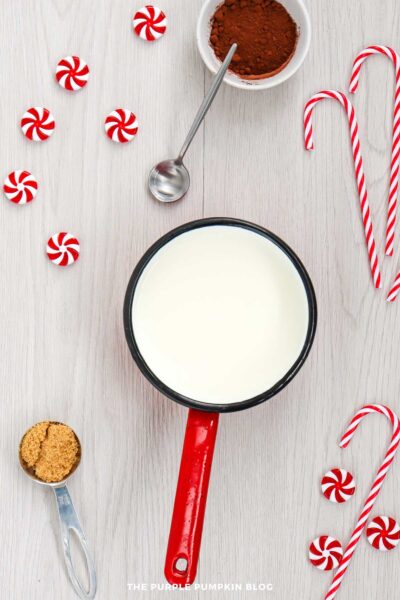 A saucepan filled with milk sits on a light wooden surface surrounded by peppermint candies, a bowl of cocoa powder with a spoon, candy canes, and a measuring spoon of brown sugar.