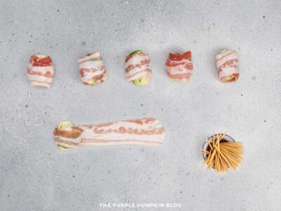 Brussels sprouts wrapped in raw bacon arranged in a row on a gray surface, with toothpicks placed to the side.