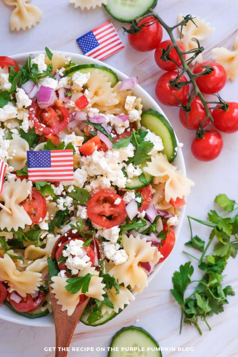 White bowl filled with bowtie pasta salad topped with cherry tomatoes, cucumber slices, red onion, parsley, crumbled feta cheese, and small American flag picks on a light marble surface.