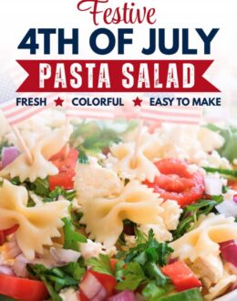 White bowl filled with bowtie pasta salad topped with cherry tomatoes, cucumber slices, red onion, parsley, crumbled feta cheese, and small American flag picks on a light marble surface.
