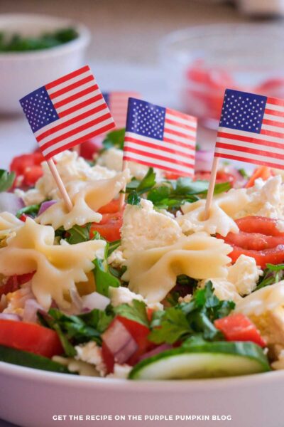 This image showcases a colorful, patriotic pasta salad decorated with small American flags, including cherry tomatoes, cucumbers, and cheese, ideal for American celebrations or summer parties.