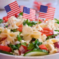 White bowl filled with bowtie pasta salad topped with cherry tomatoes, cucumber slices, red onion, parsley, crumbled feta cheese, and small American flag picks on a light marble surface.