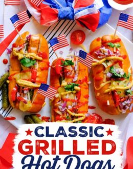Three loaded grilled hot dogs topped with cheese, pickles, diced red onion, ketchup, and mustard on a white platter with American flags and patriotic decorations. Text overlay says: "Classic Grilled Hot Dogs Perfect for 4th of July!"