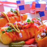 Angled view of grilled hot dogs in buns topped with ketchup, mustard, shredded cheese, diced red onion, pickles, and cilantro. Small American flags are inserted into each hot dog, with red, white, and blue party decorations in the background.