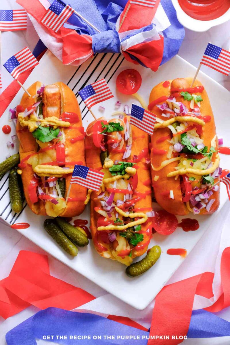Overhead view of three grilled hot dogs in toasted buns topped with ketchup, mustard, shredded cheese, diced red onion, pickles, and fresh cilantro on a white platter. Small American flags, red, white, and blue paper decorations, and cherry tomatoes surround the hot dogs.