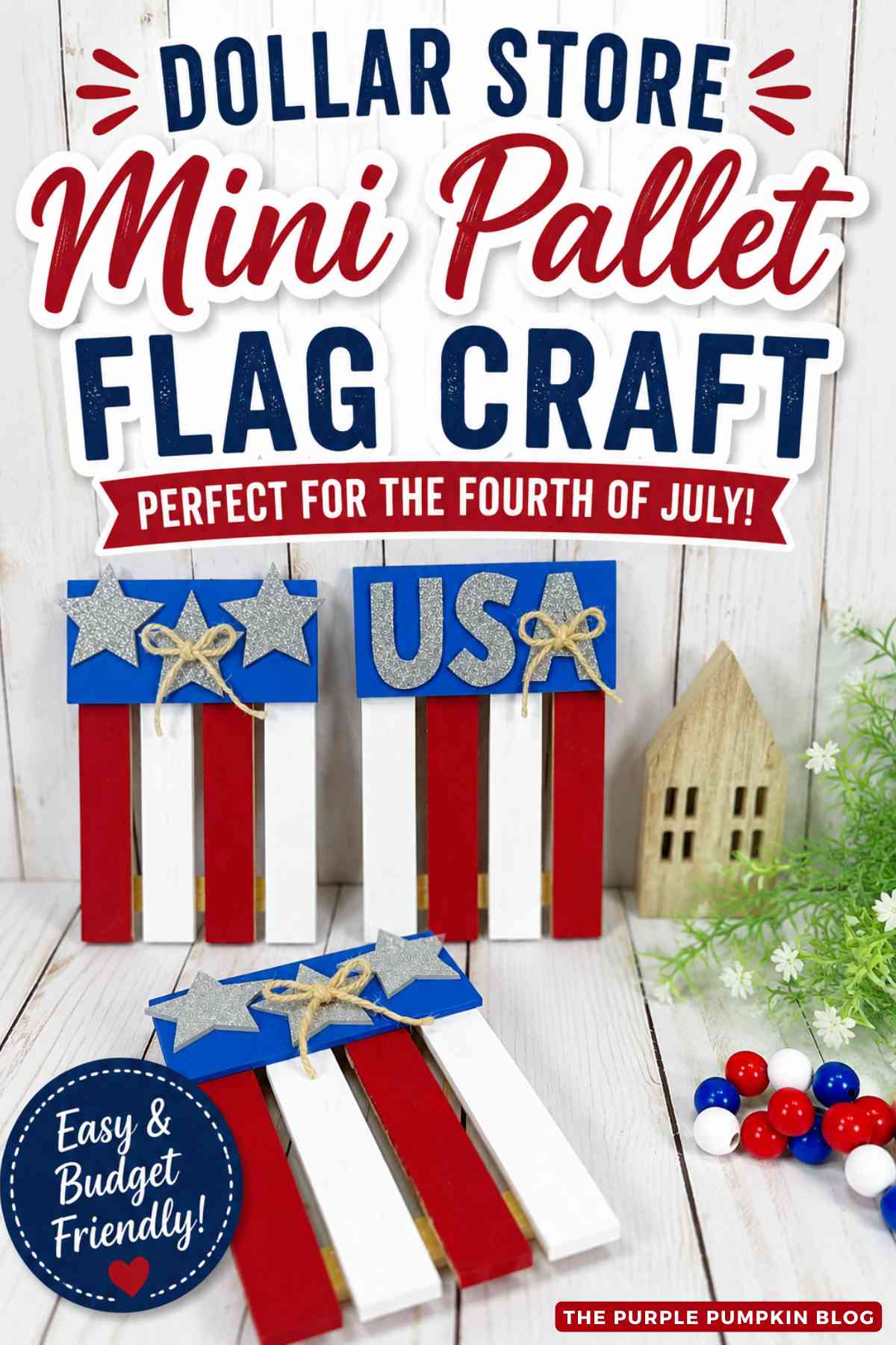 Three completed mini pallet flag crafts painted red, white, and blue. Two flags are decorated with silver glitter stars and twine bows, while the other features silver glitter letters spelling "USA." The crafts are displayed on a white wood surface with patriotic beads, greenery, and a small wooden house in the background. Text overlay says: Dollar Store Mini Pallet Flag Craft Perfect for the Fourth of July! Easy & Budget Friendly!