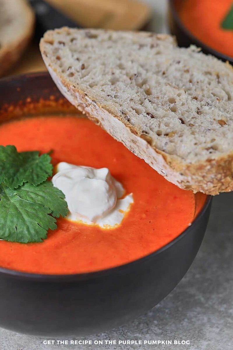 Two bowls of bright orange roasted red pepper soup topped with mascarpone cheese and parsley, served with slices of rustic seeded bread.