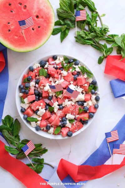 A white serving bowl filled with a watermelon salad made with juicy watermelon cubes, crumbled feta cheese, fresh blueberries, diced red onion, and mint leaves. The salad is garnished with small American flags and photographed with half a watermelon and fresh mint in the background.