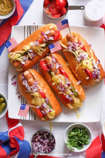 Overhead view of four grilled hot dogs in toasted buns topped with ketchup, mustard, shredded cheese, diced red onion, pickles, and fresh cilantro on a white platter. Small American flags, red, white, and blue paper decorations, and cherry tomatoes surround the hot dogs.