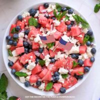 Watermelon salad with feta and blueberries served in a white bowl, featuring juicy watermelon cubes, crumbled feta cheese, fresh blueberries, diced red onion, and mint leaves. Small American flags adding a festive touch.