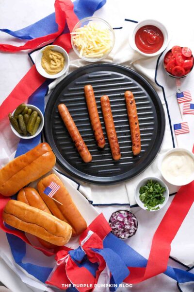 Ingredients for classic grilled hot dogs - hotdogs on a grill, buns, chopped onion, chopped herbs mayo, sliced tomatoes, ketchup, cheese, mustard, pickles.