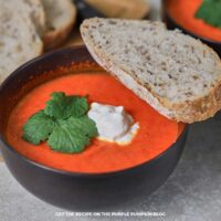 Bowl of bright orange roasted red pepper soup topped with mascarpone cheese and parsley, served with slices of rustic seeded bread.