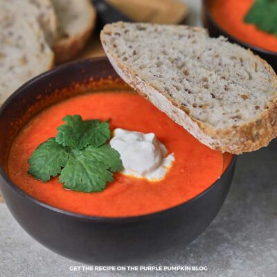 Bowl of bright orange roasted red pepper soup topped with mascarpone cheese and parsley, served with slices of rustic seeded bread.