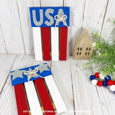 Two completed mini pallet flag crafts painted red, white, and blue. One flag is decorated with silver glitter stars and twine bows, while the other features silver glitter letters spelling "USA." The crafts are displayed on a white wood surface with patriotic beads, greenery, and a small wooden house in the background.