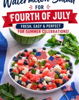 A white serving bowl filled with a watermelon salad made with juicy watermelon cubes, crumbled feta cheese, fresh blueberries, diced red onion, and mint leaves. The salad is garnished with small American flags and photographed with half a watermelon and fresh mint in the background.