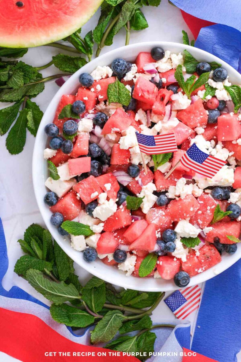 A white serving bowl filled with a watermelon salad made with juicy watermelon cubes, crumbled feta cheese, fresh blueberries, diced red onion, and mint leaves. The salad is garnished with small American flags and photographed with half a watermelon and fresh mint in the background.