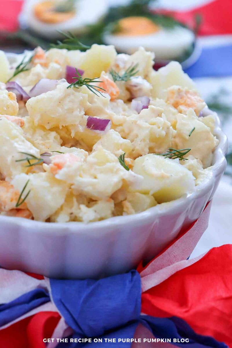 A white scalloped bowl filled with creamy potato salad made with chunks of potato, chopped hard-boiled egg, diced red onion, and fresh dill. The bowl is wrapped with red, white, and blue tissue paper, with halved hard-boiled eggs softly blurred in the patriotic background.