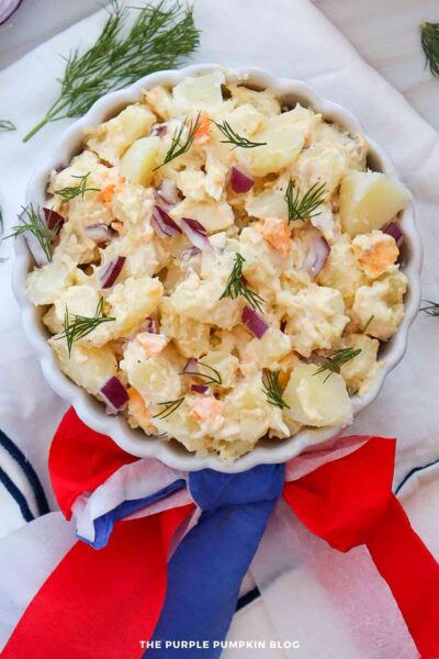 A white scalloped bowl filled with creamy potato salad made with chunks of potato, chopped hard-boiled egg, diced red onion, and fresh dill. The bowl is wrapped with red, white, and blue tissue paper.