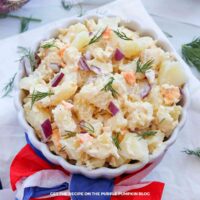 A white scalloped bowl filled with creamy potato salad made with chunks of potato, chopped hard-boiled egg, diced red onion, and fresh dill. The bowl is wrapped with red, white, and blue tissue paper.
