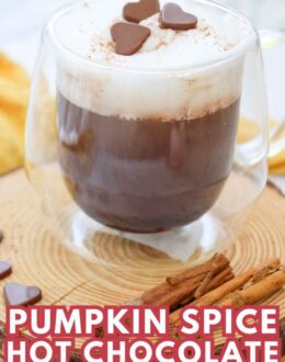 A glass mug of pumpkin spice hot chocolate topped with whipped cream and chocolate hearts, surrounded by cinnamon sticks on a wooden surface.