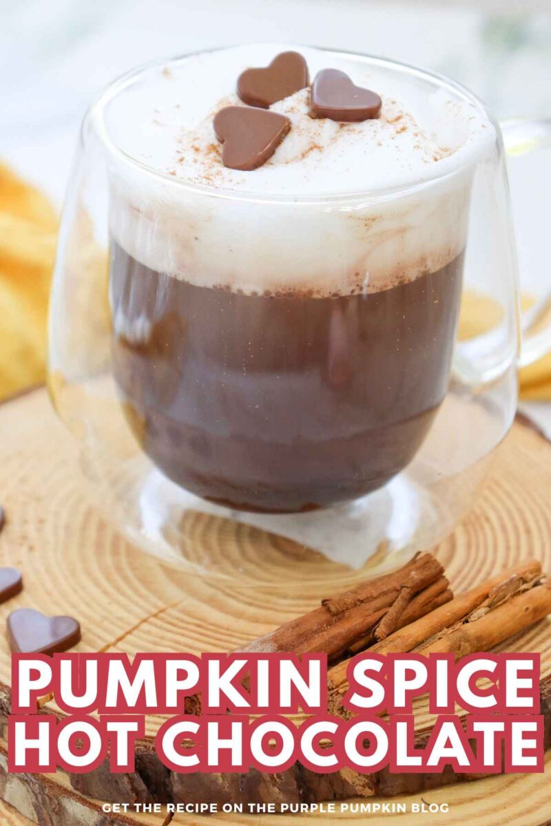 A glass mug of pumpkin spice hot chocolate topped with whipped cream and chocolate hearts, surrounded by cinnamon sticks on a wooden surface.