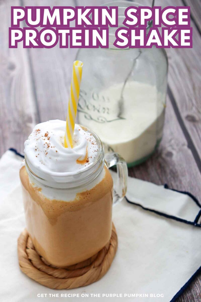 A glass jar filled with pumpkin spice protein shake topped with whipped cream and a yellow striped straw, placed on a wooden surface with a milk jug in the background.