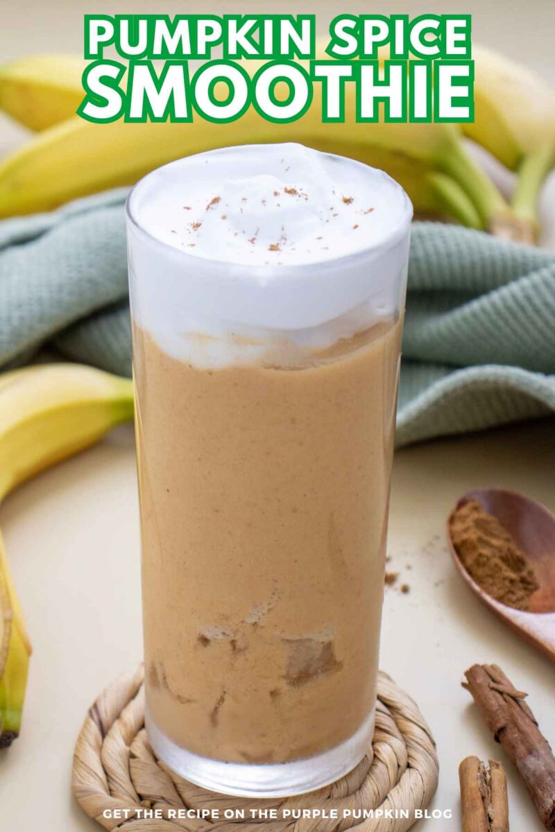 A tall glass of pumpkin spice smoothie with whipped cream and cinnamon, surrounded by bananas and cinnamon sticks on a light background.